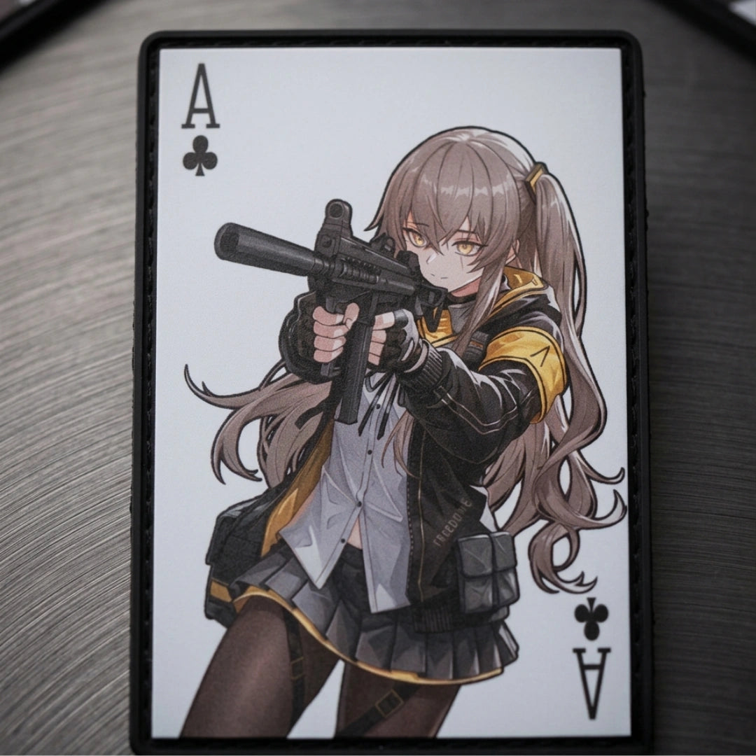A rectangular card or plaque featuring an illustrated anime-style female character with long light brown hair, aiming a black gun, with the letter A and club symbol in the corners. The card is photographed on a dark textured surface.