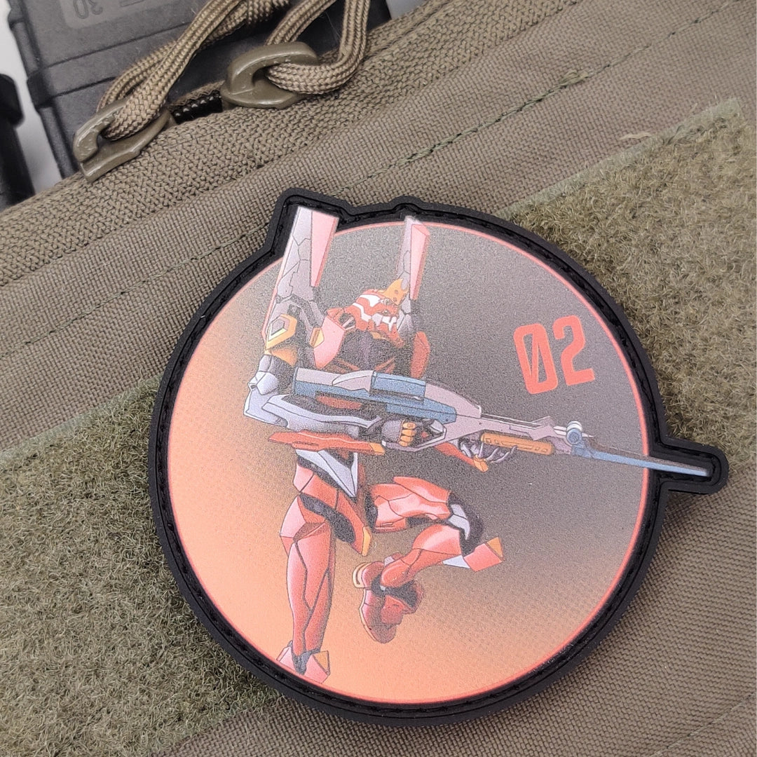 A circular tactical morale patch featuring an anime-style mecha character in red and orange armor holding a rifle, with the number '02' printed in orange on a peach-to-gray gradient background. The patch is shown attached to a tan canvas strap.