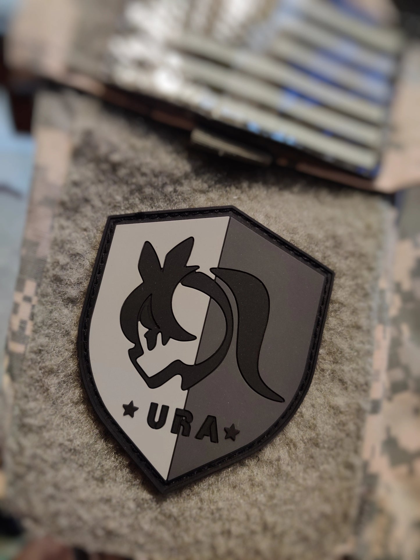 Shield-shaped logo with Umamusume Racing Association emblem and 'URA' text on a gray camoflauge texture background