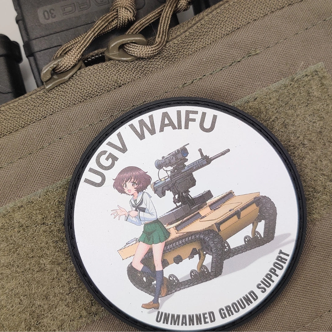 A circular tactical patch featuring an anime-style illustration of a girl next to a UGV tank vehicle, with the text 'UGV WAifu' and 'Unmanned Ground Support'. The patch is shown pinned to a tan tactical fabric background.