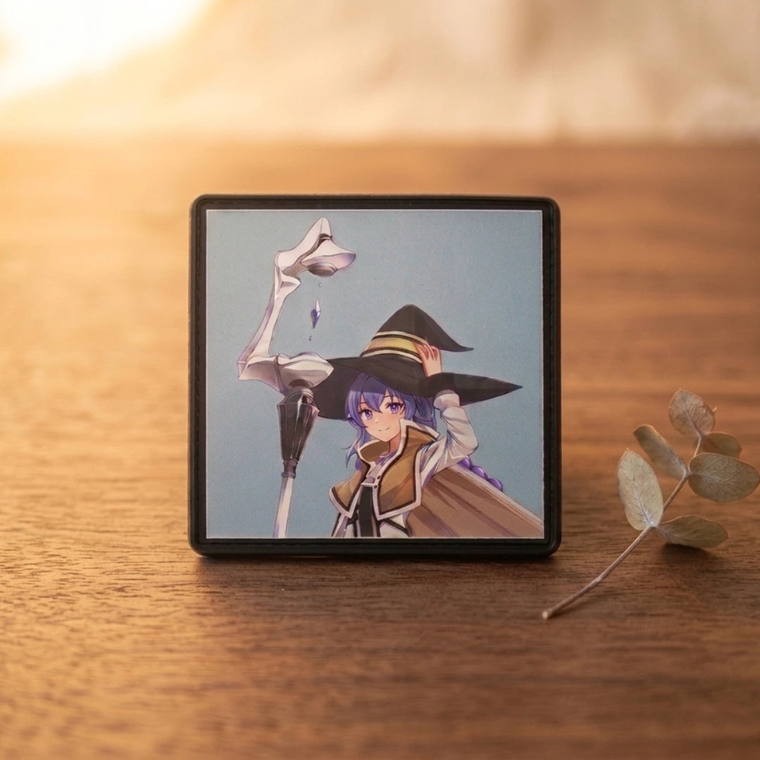 A square black-framed picture frame or small photo print displaying an anime-style character with purple hair, a large black hat, and a white bird, placed on a wooden surface with a blurred warm background and a small eucalyptus sprig nearby.
