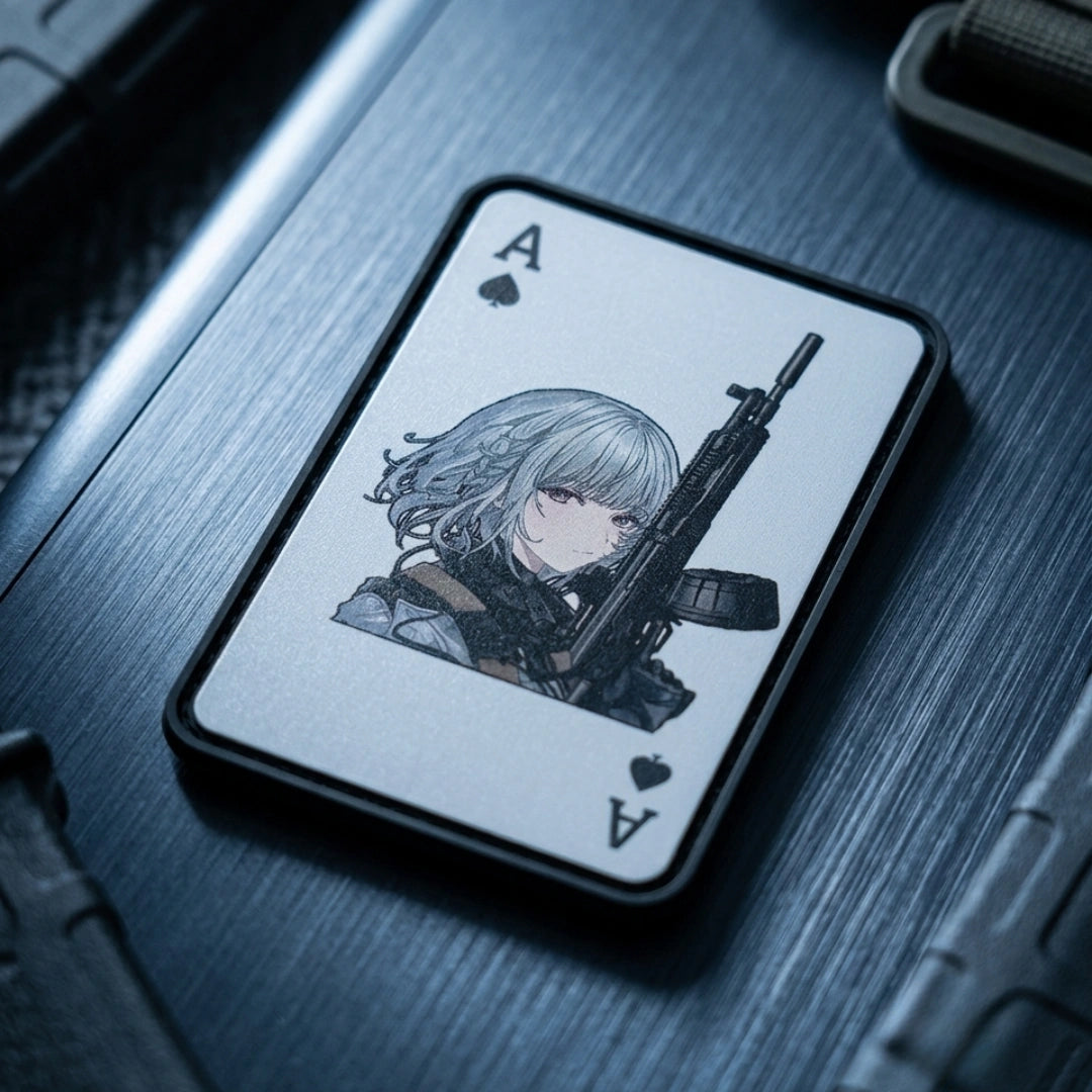 A rectangular card or deck box featuring an illustrated anime-style character with silver hair holding a rifle, shown on a dark metallic surface. The card design includes black spade and heart symbols with the letter A in the corners.