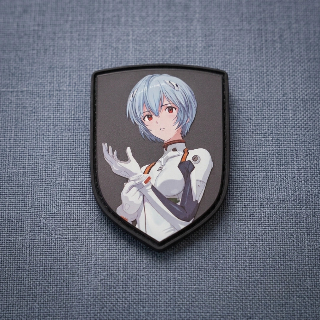 A shield-shaped embroidered patch featuring an anime-style character with short light blue hair, red eyes, and a white outfit, displayed on a textured blue-gray fabric background.