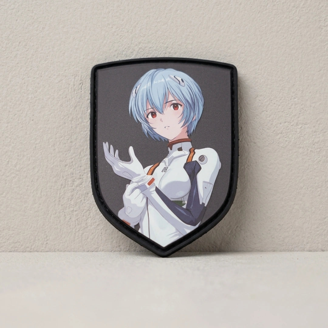 A shield-shaped patch featuring an anime character with short light blue hair and red eyes, wearing a white and black outfit, displayed against a textured beige wall.