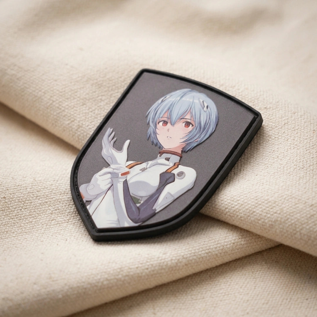 A shield-shaped patch featuring an anime-style character with short silver hair and a white and gray outfit, resting on a beige textured fabric background.