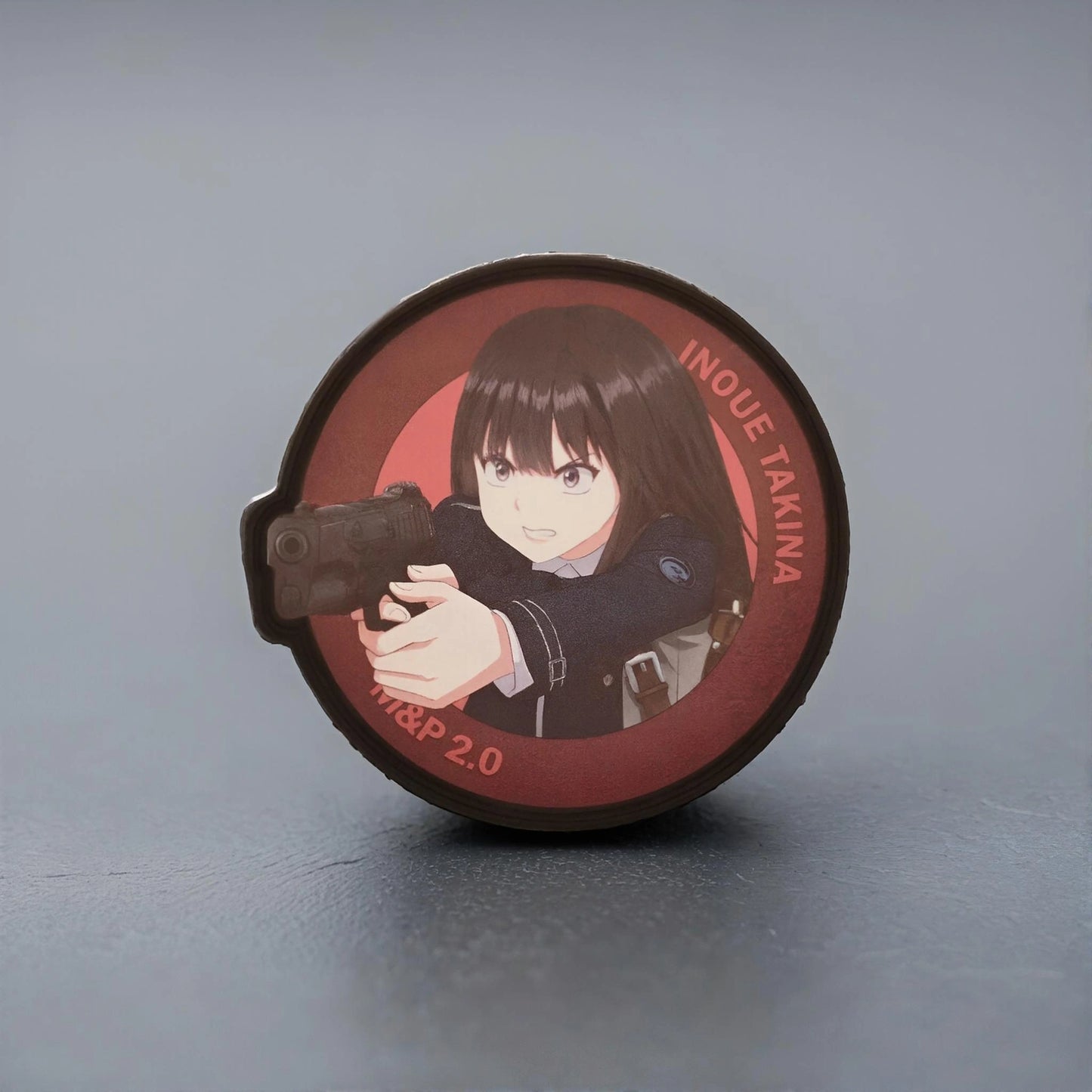 Circular product with an illustration of Takina Inoue from the anime Lycoris Recoil holding a gun on a gray background