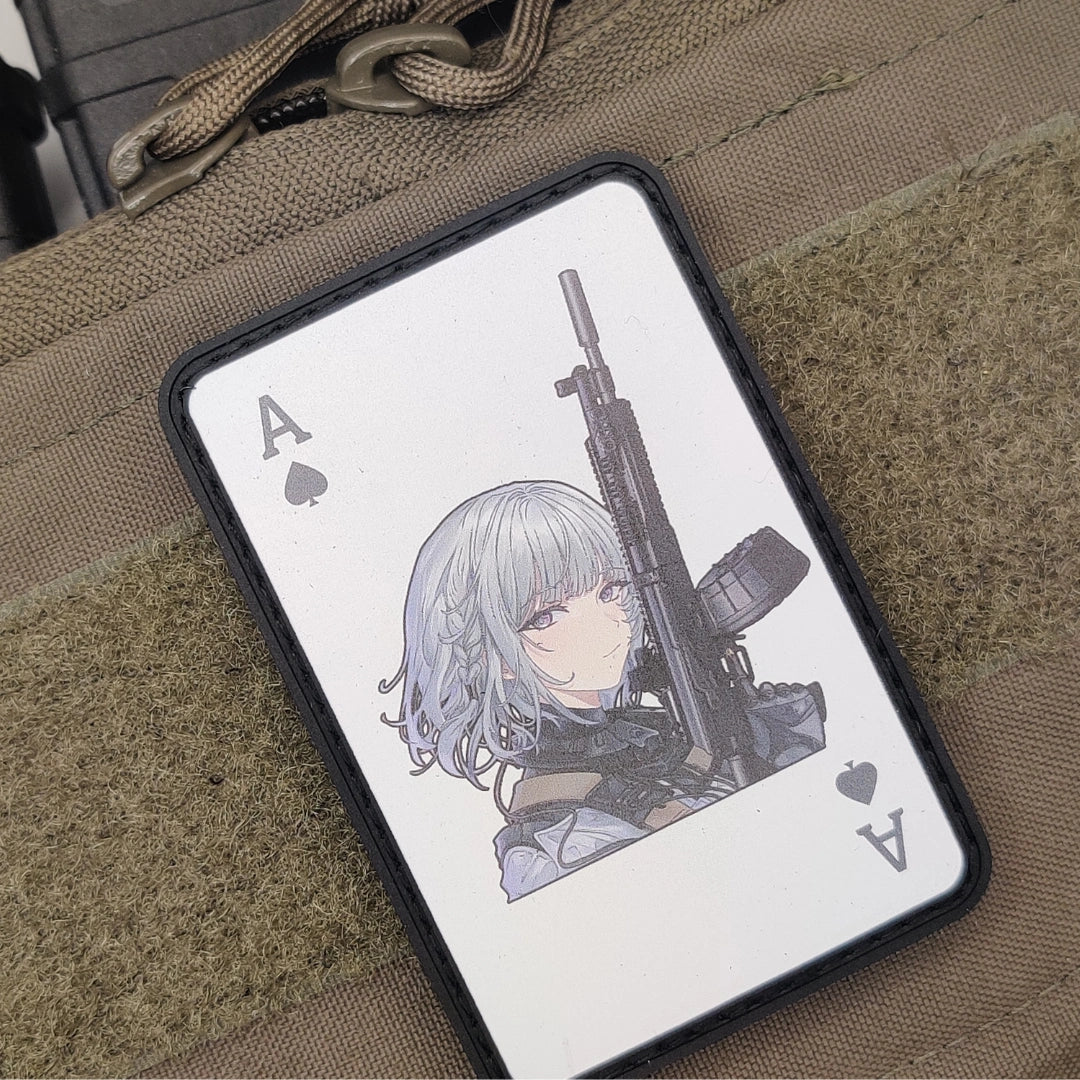 A rectangular black-framed card case or phone skin featuring an illustrated anime-style character with silver hair holding a rifle, styled on a tan tactical fabric surface.