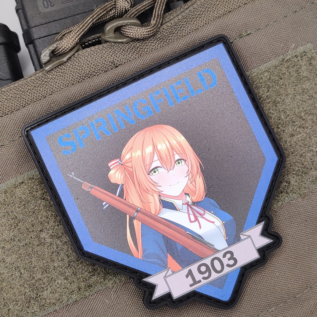 A shield-shaped tactical patch featuring an anime-style illustration of a girl with orange hair holding a rifle, with the text 'SPRINGFIELD' at the top and '1903' on a banner at the bottom, shown attached to a tan fabric surface.