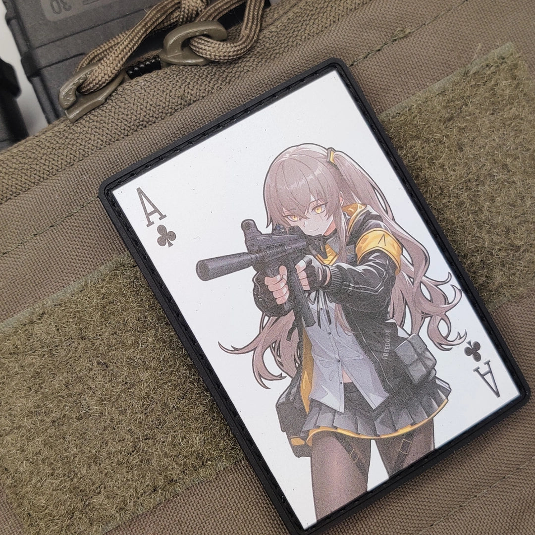 A rectangular card or sticker featuring an anime-style character holding a gun, printed on a black-bordered card with Ace of Clubs suit marks, photographed on a tan textured fabric surface.
