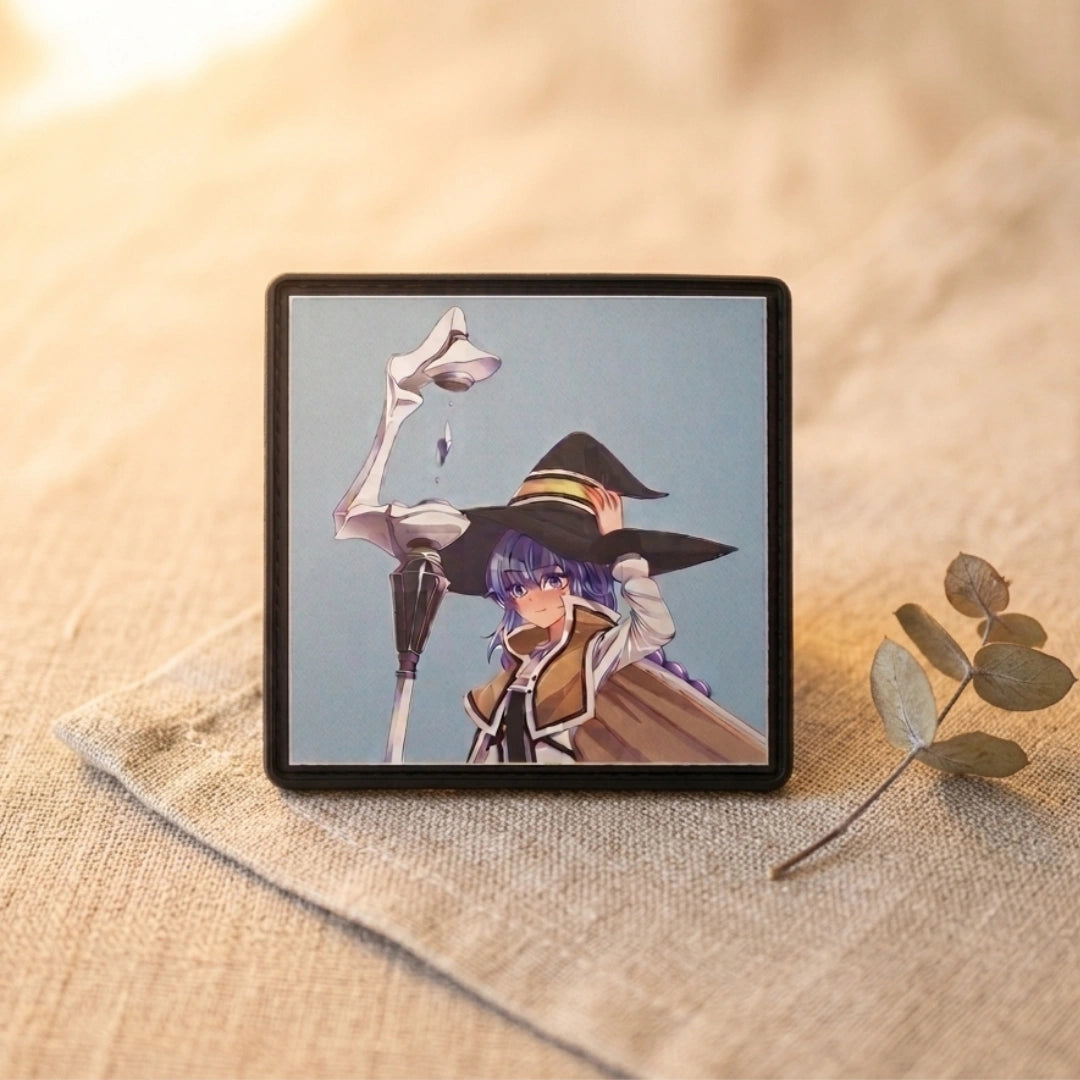 A square black-framed art print or small picture box featuring an anime-style character in a witch hat, placed on a beige fabric surface with a dried leaf nearby.