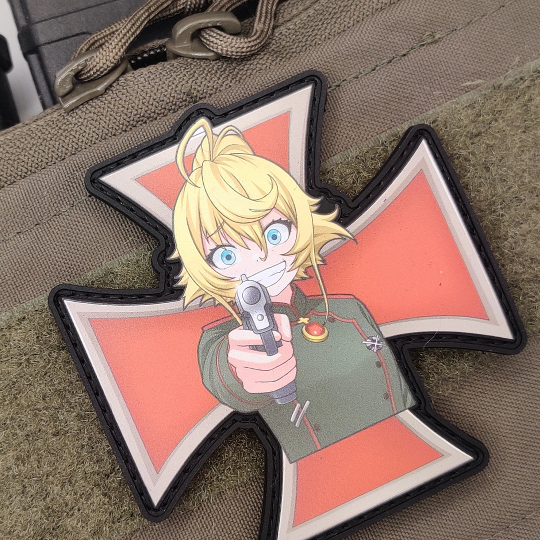 A tactical patch shaped like an Iron Cross, featuring an anime character with blonde hair and blue eyes holding a gun, set against a coral background with a black and white border, displayed on a tan canvas surface.