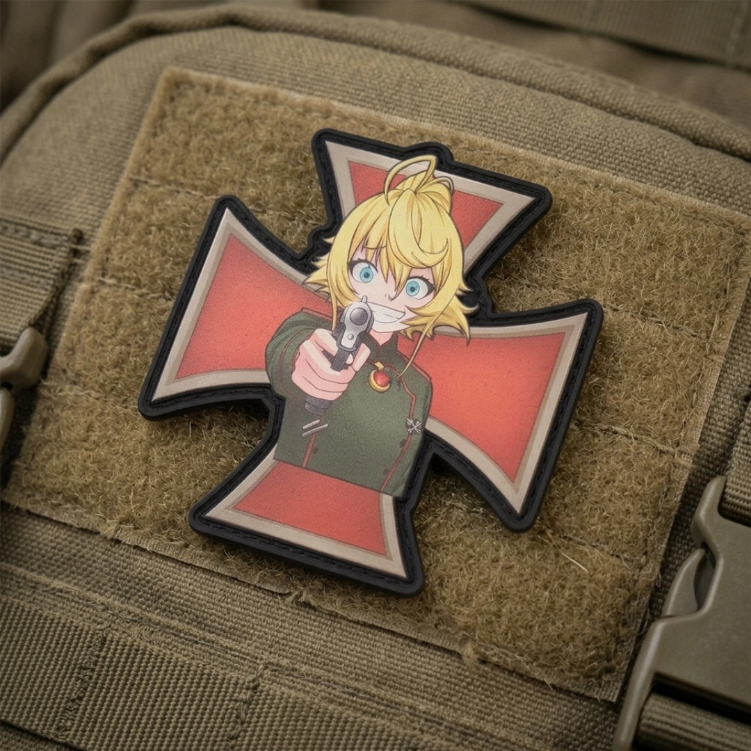 An anime-themed tactical patch featuring a blonde character pointing a gun, set on a red and black Iron Cross background, shown attached to a tan tactical backpack.